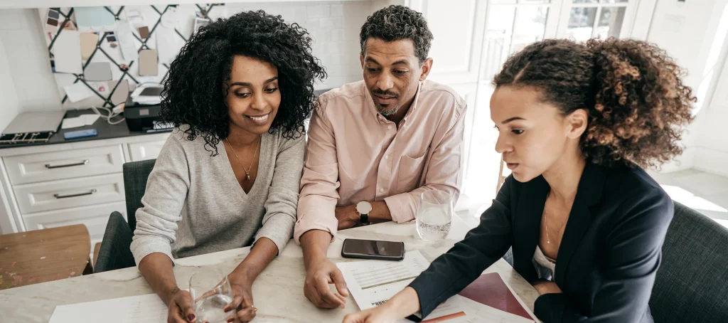 A realtor sits at a desk with two couples, presenting them with various property listings.