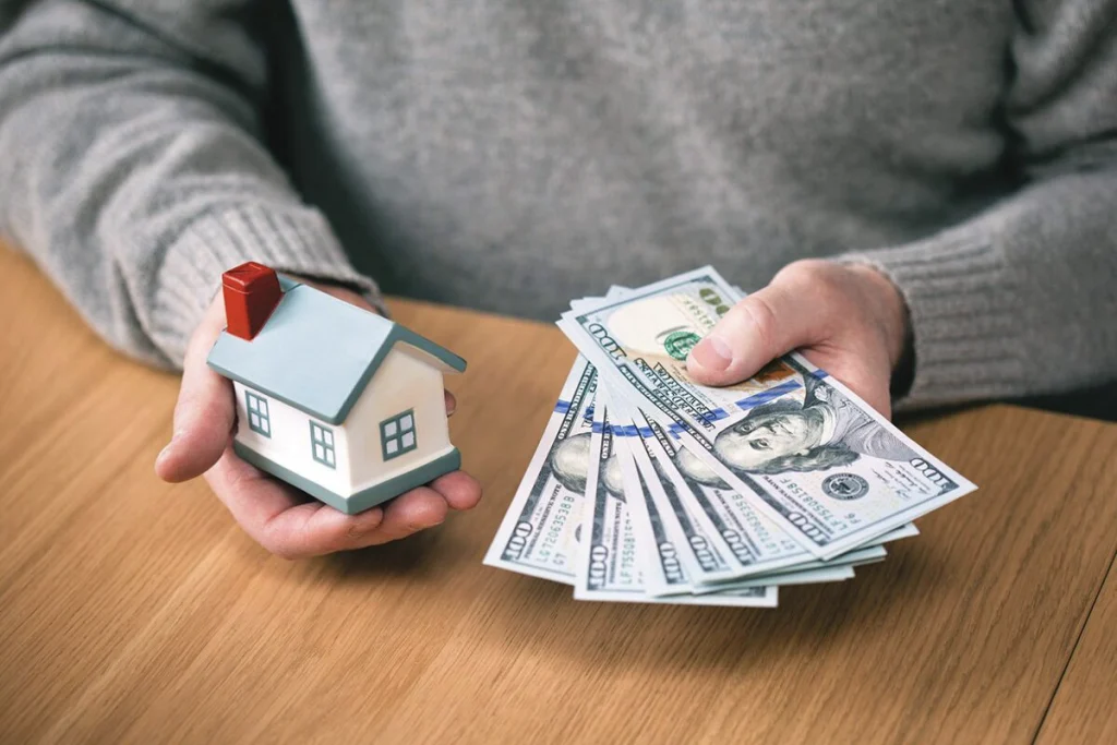 An image depicting a man's hands resting on a table in his house, with one hand holding a house emblem and the other hand clutching cash.
