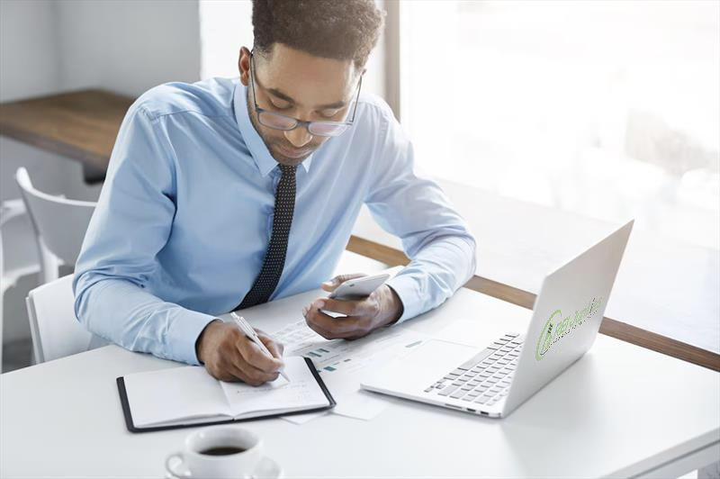 a picture of a Man writing at notebook on his desk, and a laptop in front of him.