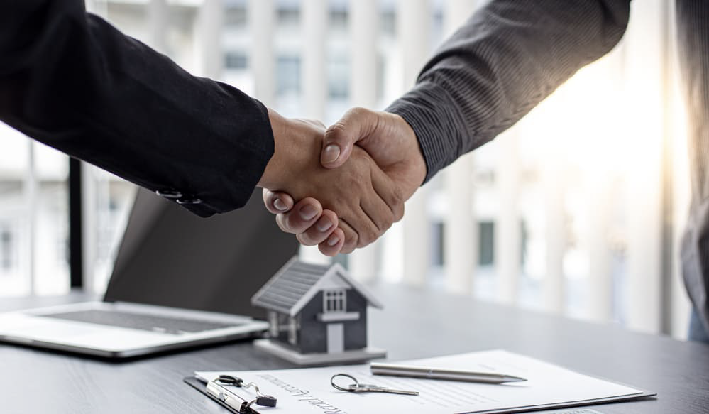 An image displays a check hand above a desk where a contract for a closing house, featuring the house emblem, and a laptop are situated.