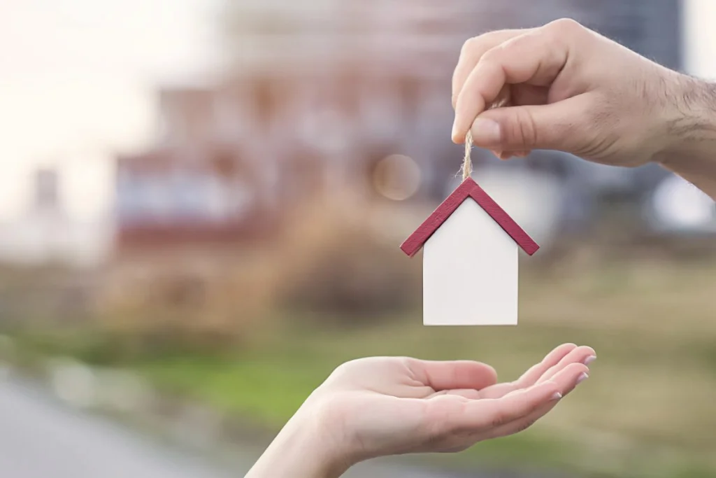 An image portrays two hands, with one hand presenting a figurine resembling a house, indicative of a sale.